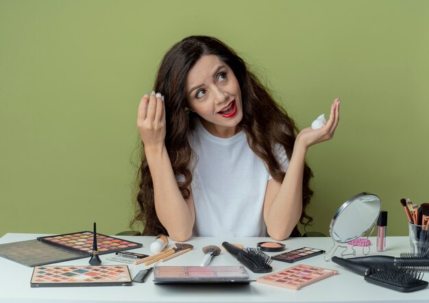 Impressed young pretty girl sitting at makeup table with makeup tools applying hair mousse on hair and looking at side isolated on olive green background