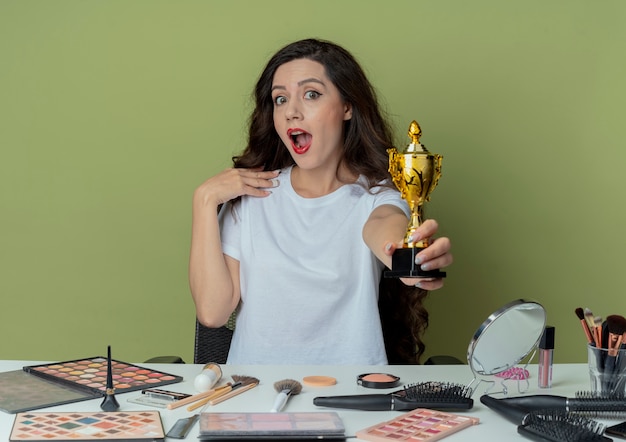 Free photo impressed young pretty girl sitting at makeup table with makeup tools stretching out winner cup towards camera and touching chin isolated on olive green background