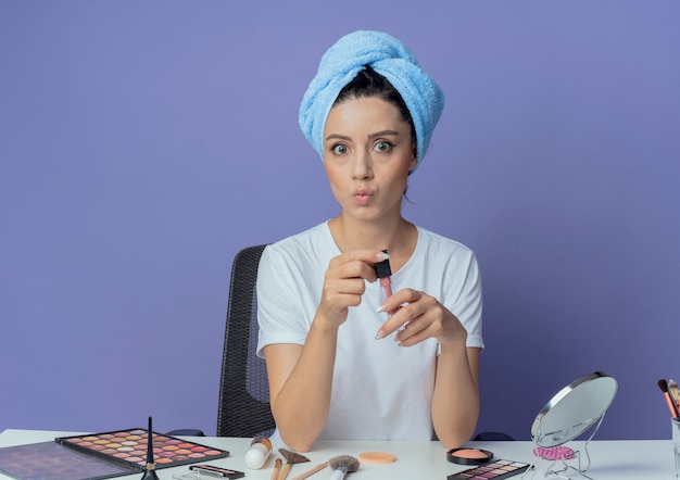 Free photo impressed young pretty girl sitting at makeup table with makeup tools and with bath towel on head holding lipgloss