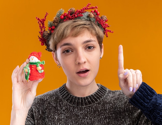 Free photo impressed young pretty girl wearing christmas head wreath holding small christmas snowman statue looking at camera pointing up isolated on orange background