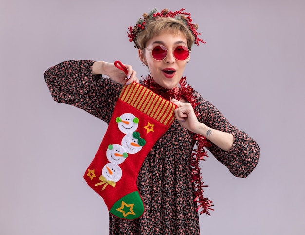 Free Photo impressed young pretty girl wearing christmas head wreath and tinsel garland around neck with glasses holding christmas stocking looking at camera isolated on white background