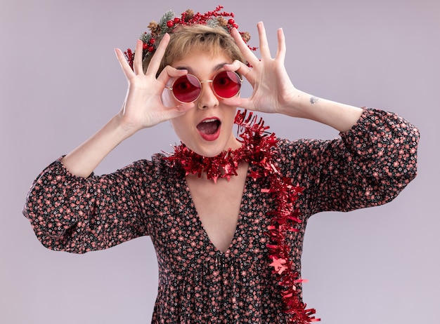 Impressed young pretty girl wearing christmas head wreath and tinsel garland around neck with glasses looking at camera grabbing glasses doing look gesture isolated on white background