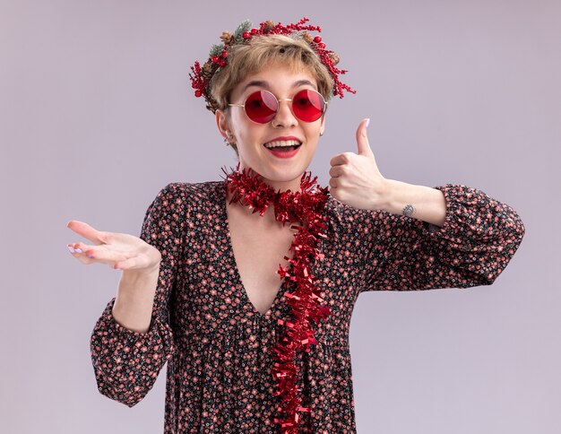 Impressed young pretty girl wearing christmas head wreath and tinsel garland around neck with glasses looking at camera showing empty hand and thumb up isolated on white background