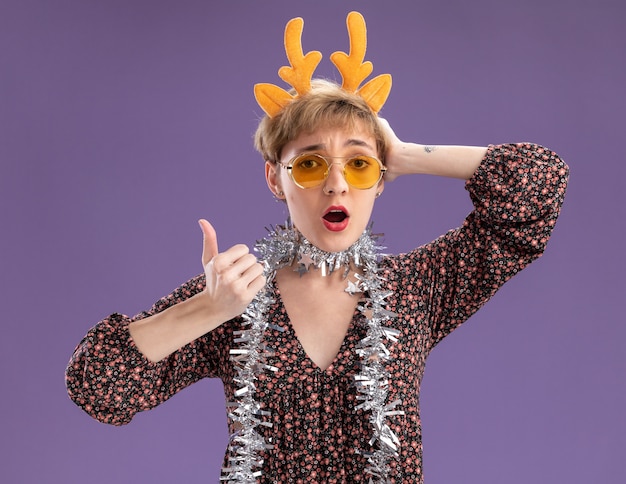 Free Photo impressed young pretty girl wearing reindeer antlers headband and tinsel garland around neck with glasses looking at camera keeping hand on head showing thumb up isolated on purple background