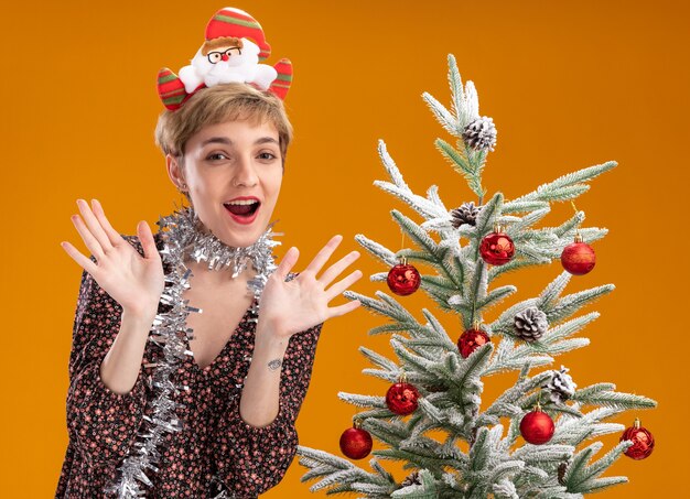 Impressed young pretty girl wearing santa claus headband and tinsel garland around neck standing near decorated christmas tree showing empty hands isolated on orange background