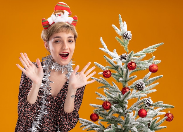 Impressed young pretty girl wearing santa claus headband and tinsel garland around neck standing near decorated christmas tree showing empty hands isolated on orange wall