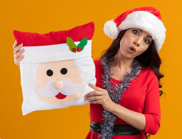 Free Photo impressed young pretty girl wearing santa hat and tinsel garland around neck holding santa claus pillow looking at camera isolated on orange background