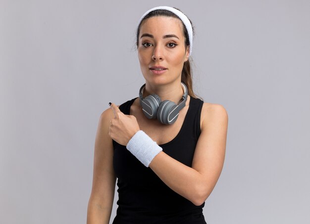 Impressed young pretty sporty girl wearing headband and wristbands with headphones around neck pointing behind 