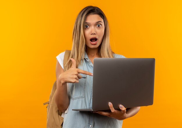 Impressed young pretty student girl wearing back bag holding and pointing at laptop isolated on orange