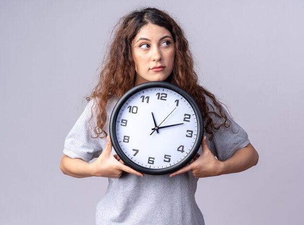 Impressed young pretty woman holding clock looking at side isolated on white wall