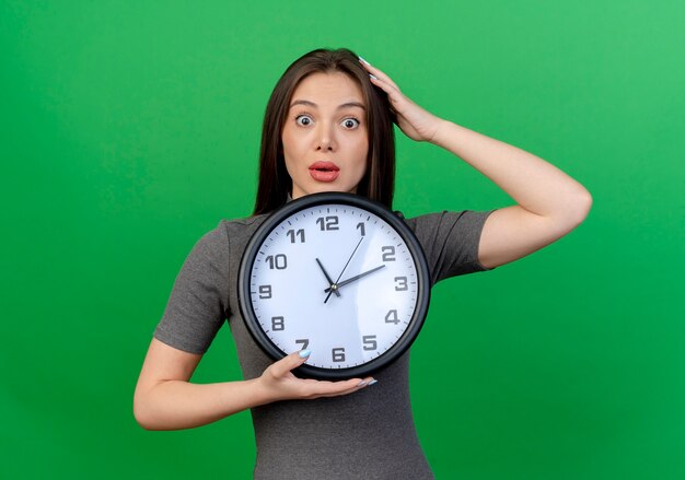 Impressed young pretty woman holding clock and putting hand on head isolated on green background