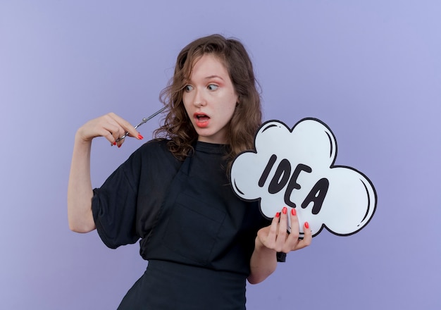 Impressed young slavic female barber wearing uniform holding scissors and idea bubble looking at scissors isolated on purple background