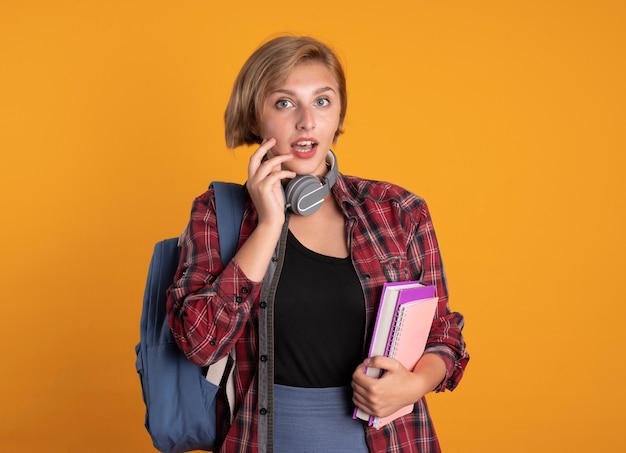 Impressed young slavic student girl with headphones wearing backpack holds book and notebook 