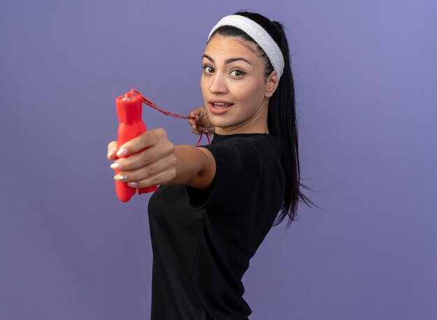 Impressed young sporty woman wearing headband and wristbands standing in profile view pulling jump rope stretching it out towards front looking at front isolated on purple wall