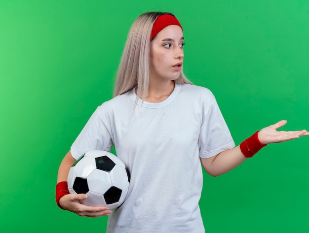 Impressed young sporty woman with braces wearing headband and wristbands holds ball looking and pointing at side isolated on green wall