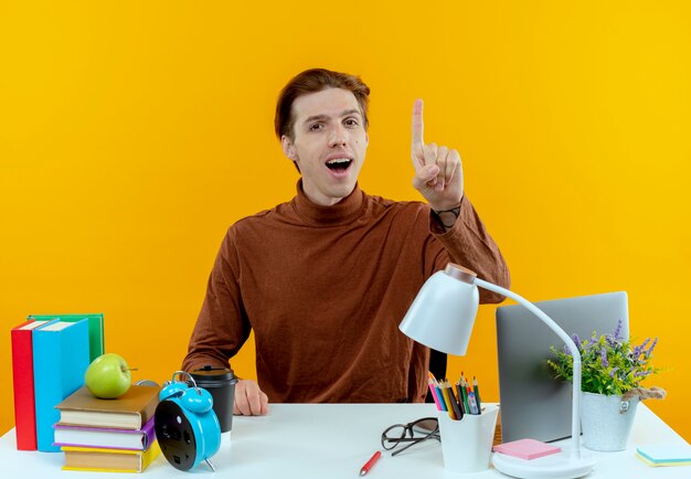 Impressed young student boy sitting at desk with school tools points at up 