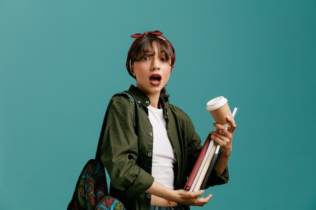 Free photo impressed young student girl wearing bandana and backpack holding note pads and paper coffee cup looking at camera isolated on blue background