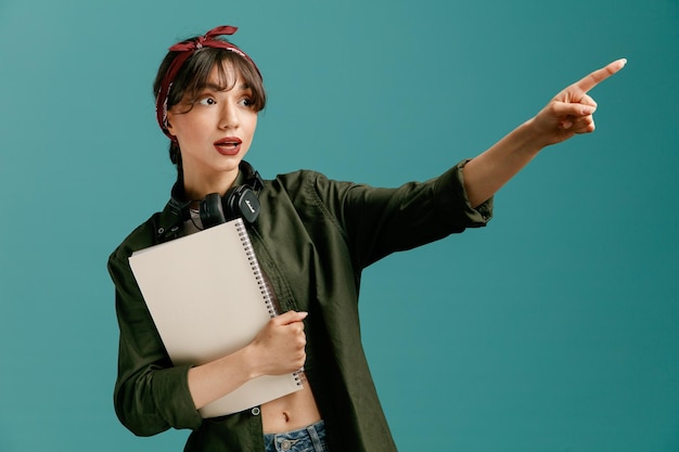 Free photo impressed young student girl wearing bandana and headphones around neck holding large note pad looking at side pointing to side isolated on blue background