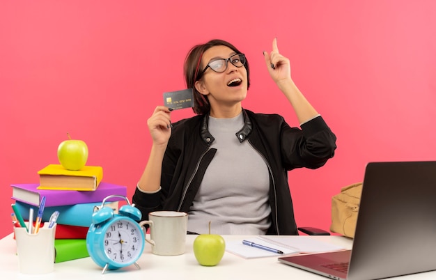 Impressed young student girl wearing glasses sitting at desk holding credit card raising finger isolated on pink