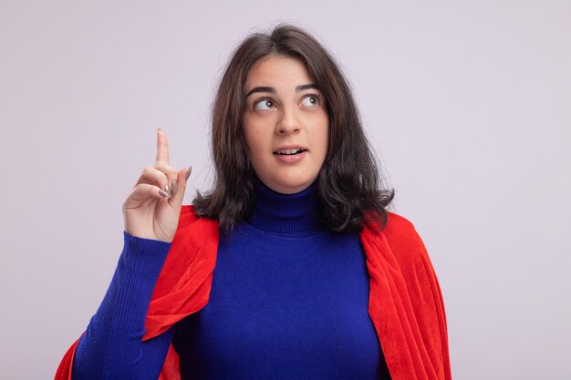 Impressed young superhero woman in red cape looking at side pointing up isolated on white wall