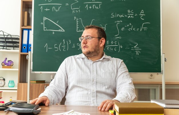 impressed young teacher wearing glasses sitting at desk with school supplies in classroom keeping hands on desk looking at side