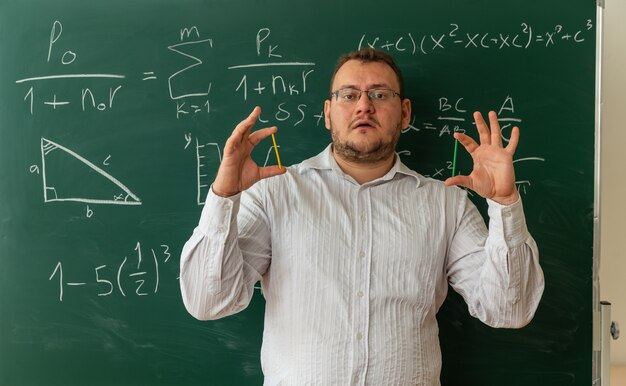 impressed young teacher wearing glasses standing in front of chalkboard in classroom looking at front showing counting sticks