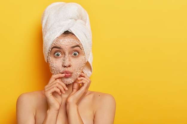 Free photo impressed young woman keeps lips rounded, has wide opened eyes, applies sea salt scrub, wears wrapped towel on head, has delicate soft skin, models indoor. people, spa, peeling, freshness concept