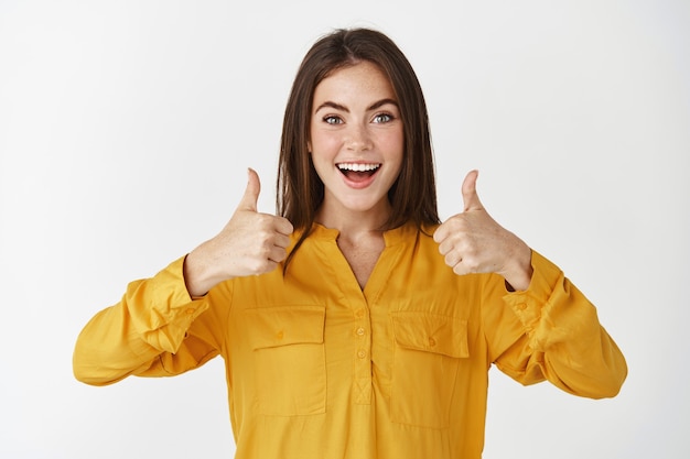 Impressed young woman showing thumbs up and smiling amazed, praising something cool, standing over white wall