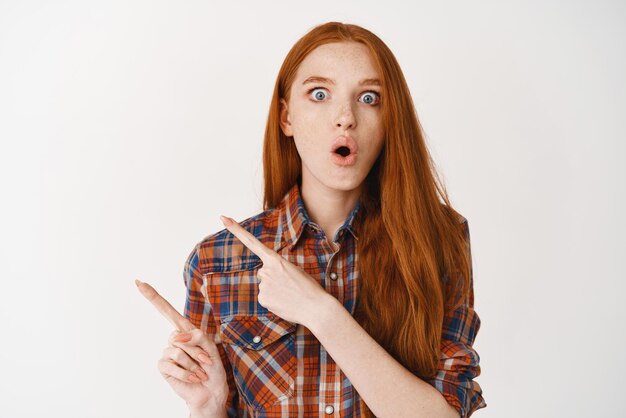 Impressed young woman with red long hair and pale skin staring amazed and pointing fingers left at advertisement standing over white background