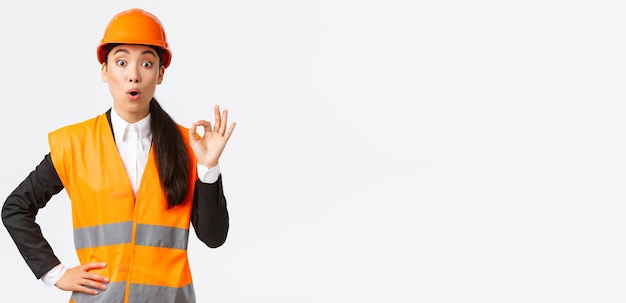 Imressed and surprised asian female construction manager pleased with amazing quality standing in safety helmet and reflective jacket showing okay gesture and say wow white background