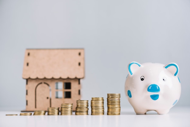 Increasing stacked coins; piggybank and house model on white reflective desk