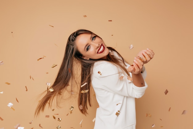 Free photo incredible female model with lovely smile and long light-brown hair wearing white jacket posing on beige wall with confeti and preparing for birthday party