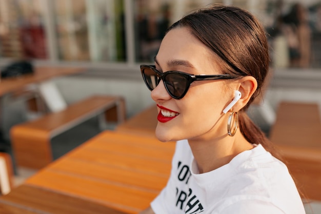 incredible woman with widely smile and red lips wearing dark sunglasses and white tshirt sitting on summer terrace