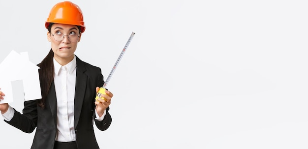 Indecisive asian female architect construction engineer in helmet and business suit holding house maket and tape measure biting lip unsure looking away thoughtful searching solution