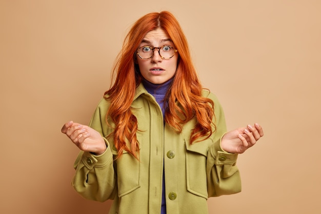 Free Photo indecisive confused redhead woman spreads palms sideways and feels confused stares with shocked expression wears fashionable outerwear.