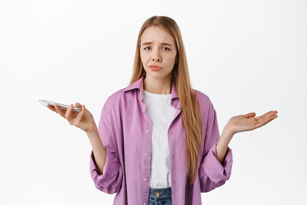 Free Photo indecisive cute girl holding smartphone spread hands sideways and shrugging confused dont know nothing what to do standing over white background