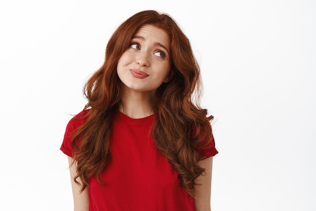 Indecisive cute redhead woman looking aside avoid eye contact and shrugging unsure hesitating standing coy in tshirt against white background