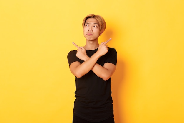 Indecisive handsome asian guy looking confused, pointing fingers sideways, standing yellow wall