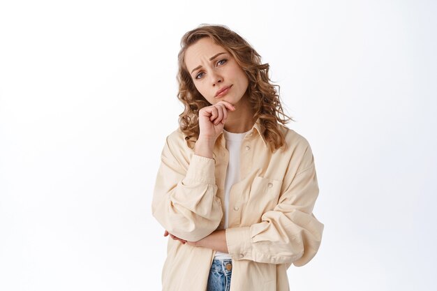 Indecisive lady looking at front thoughtful, making decision, frowning and staring pensive, standing in modern clothes against white wall