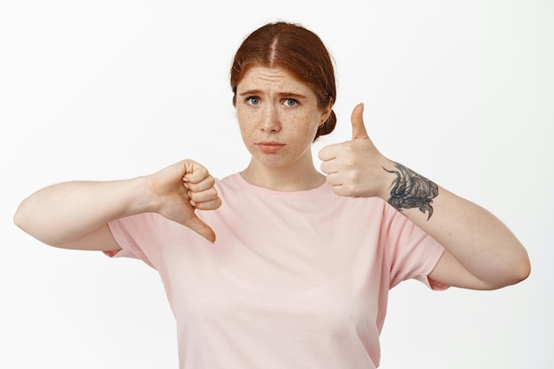 Indecisive redhead girl showing thumbs up, thumb down and look perplexed, judging average not bad result, hesitating to rate something, standing over white background