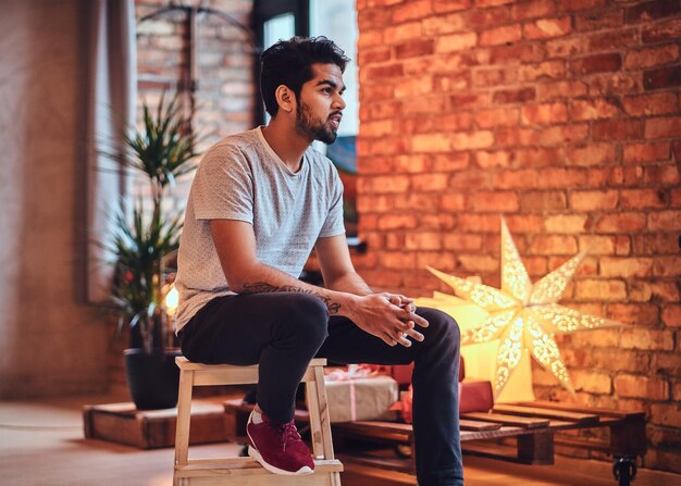 Indian bearded hipster male with tattoos on the arm in a loft room with Christmas decoration.