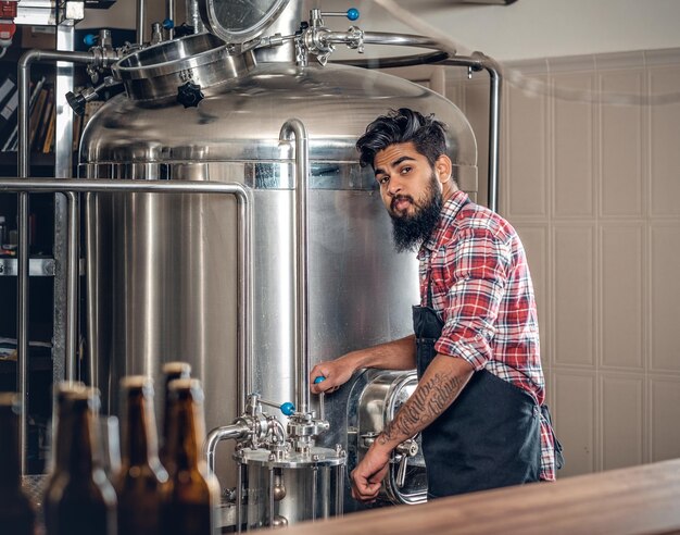 Indian bearded tattooed hipster male manufacturer presenting craft beer in the microbrewery.