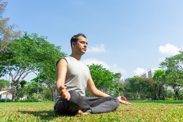 Indian man meditating in lotus pose outdoors on summer lawn with trees in background.