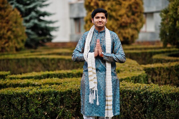 Indian man wear on traditional clothes with white scarf posed outdoor against green bushes at park show namaste hands sign