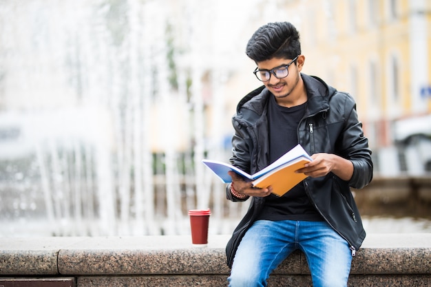 Free photo indian student man holding a pile of books sitting near fountain on the street