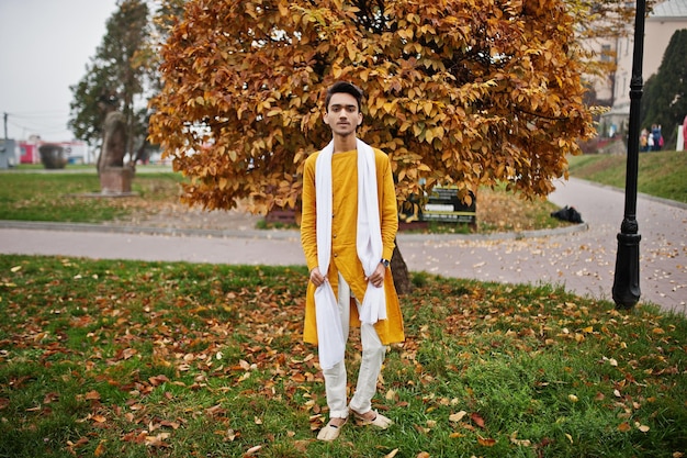 Indian stylish man in yellow traditional clothes with white scarf posed outdoor against autumn leaves