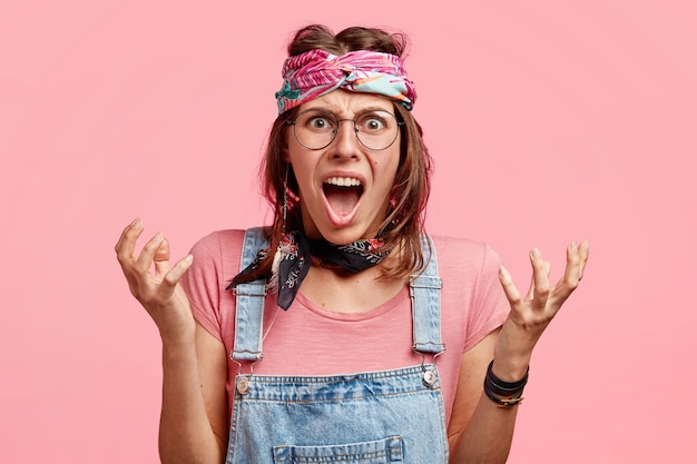 Free Photo indignant angry hippy woman gestures with hands, exclaims furiously, expresses negative emotions, dressed in fashionable overalls and headband, poses against pink wall