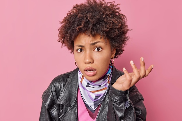 Free photo indignant curly haired young woman raises palm and looks displeased shrugs shoulders with bewilderment wears black leather jacket isolated over pink background. human reactions concept. so what