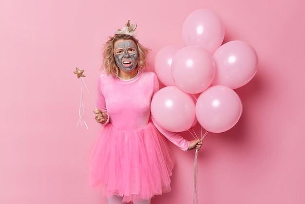 Indignant discontent young woman looks puzzled applies nourishing clay mask on face hears unpleasant news prepares for party and celebration isolated over pink background holds inflated balloons