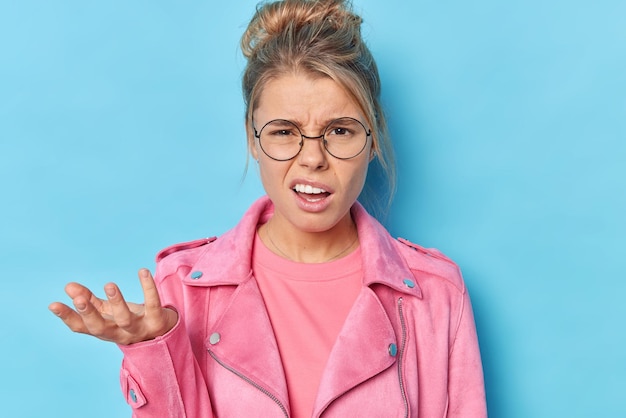 Free photo indignant discontent young woman shrugs shouders says so what frowns face cannot understand whats happening dressed in fashionable pink jacket round spectacles isolated over blue background.